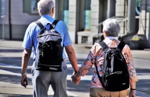 Senior couple holding hands and wearing backpacks while walking.